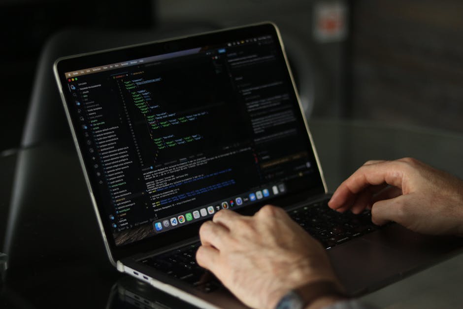 A close-up view of a modern, slim silver laptop displaying lines of colorful programming code on its screen, positioned on a light wooden desk. Next to the laptop, there is an open, spiral-bound noteb